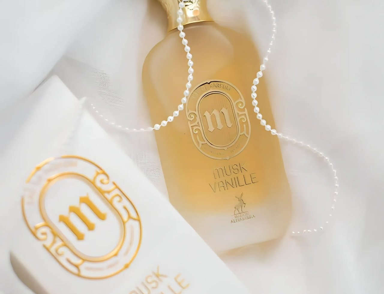 Maison Alhambra Musk Vanille by Maison Alhambra 100ml EDP — Botol minyak wangi kaca beku dengan penutup emas, kalung mutiara dililit padanya, dan kotak persembahan putih serta emas yang sepadan.