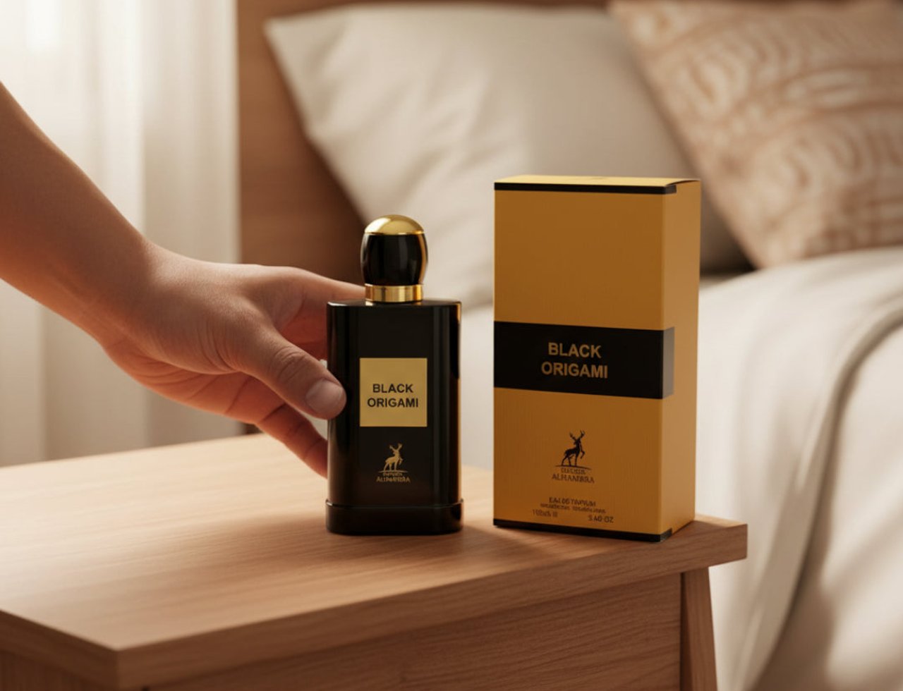 Maison Alhambra Black Origami — Tangan memegang botol minyak wangi hitam berpenutup emas, diletakkan di atas meja sisi katil kayu bersebelahan kotak emas dan hitamnya. Katil kelihatan di latar belakang.