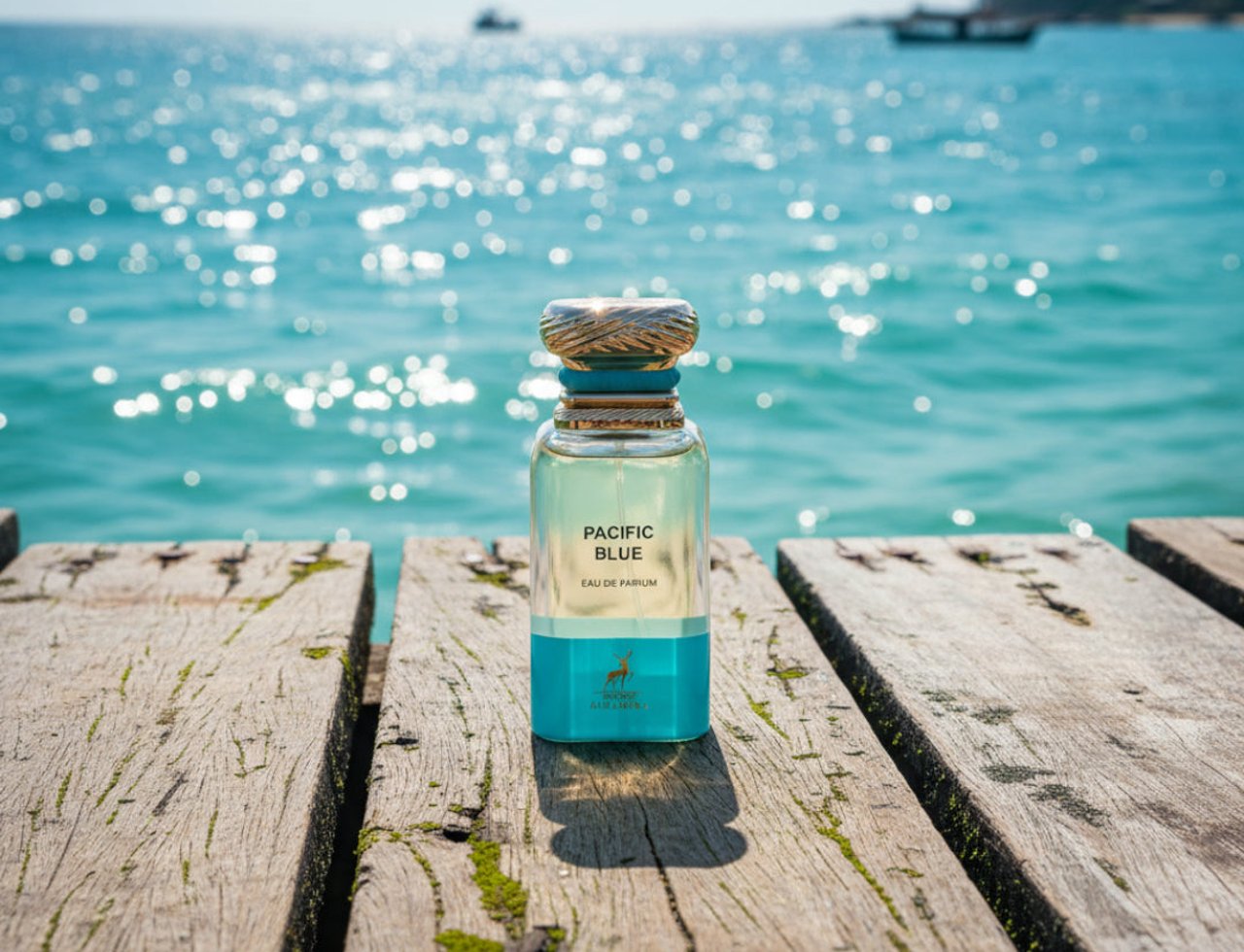 Maison Alhambra Pacific Blue by Maison Alhambra 80ml Man — A clear glass perfume bottle with a gold textured cap sits on a weathered wooden pier, with a sparkling turquoise ocean and distant boats in the background.