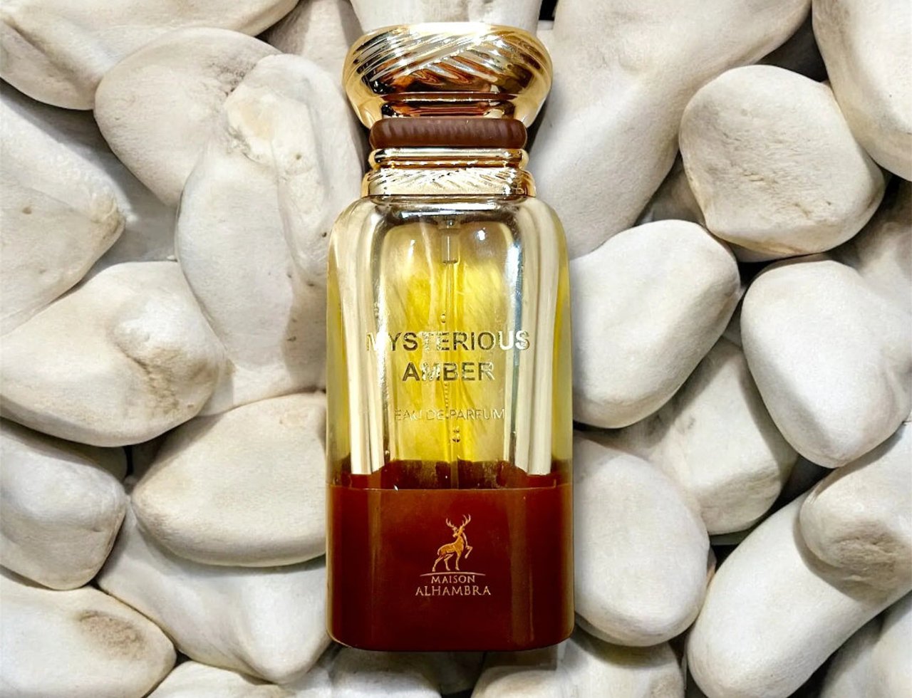 Maison Alhambra Mysterious Amber by Maison Alhambra 80ml — A clear glass perfume bottle with gold cap and amber liquid, set against a backdrop of smooth white stones.