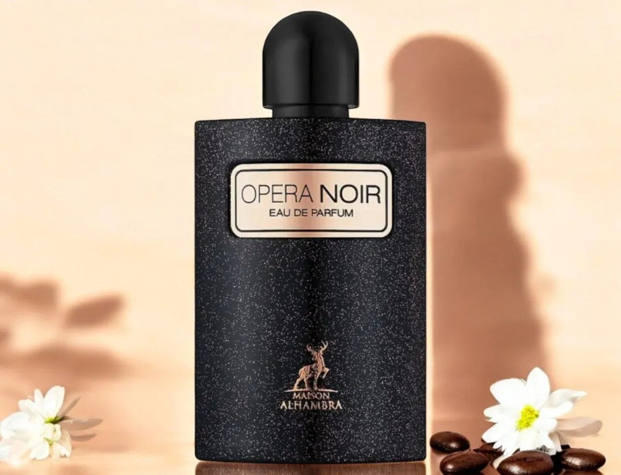 Maison Alhambra Opera Noir by Maison Alhambra 100ml EDP — Black textured bottle with gold label, deer logo, and black cap, set against a soft peach background with shadows and white flowers.