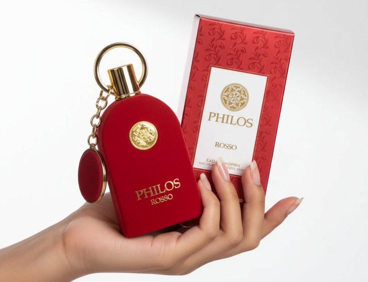 Maison Alhambra Philos Rosso — Red velvet perfume bottle with gold accents and keychain, held by a hand next to its red patterned box.