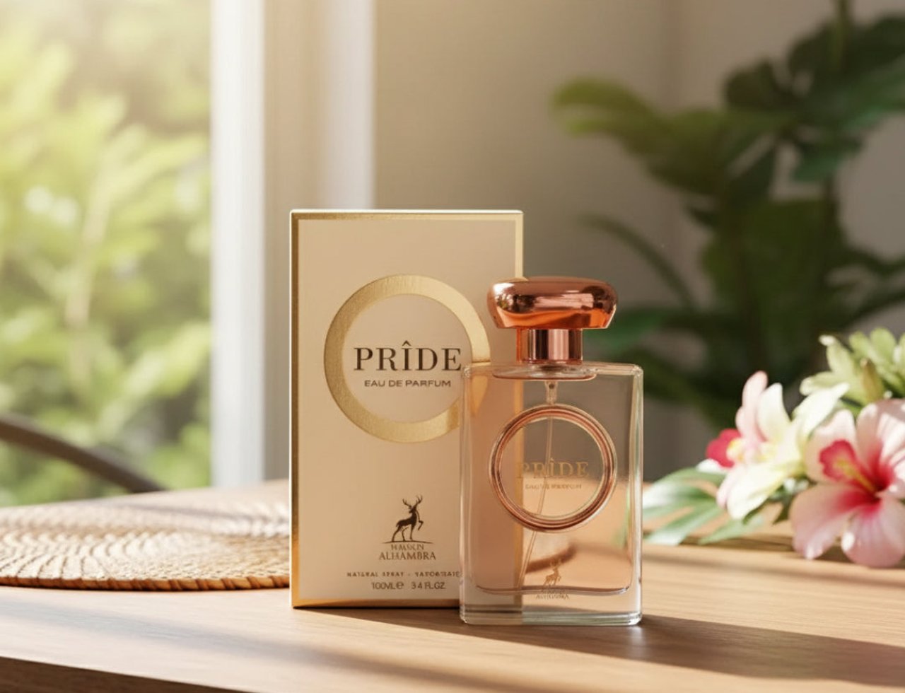 Maison Alhambra Pride by Maison Alhambra 100ml EDP Women — A clear glass perfume bottle with a rose gold cap and a circular rose gold accent on the bottle, next to its gold box, on a wooden table with a blurred green background and flowers.