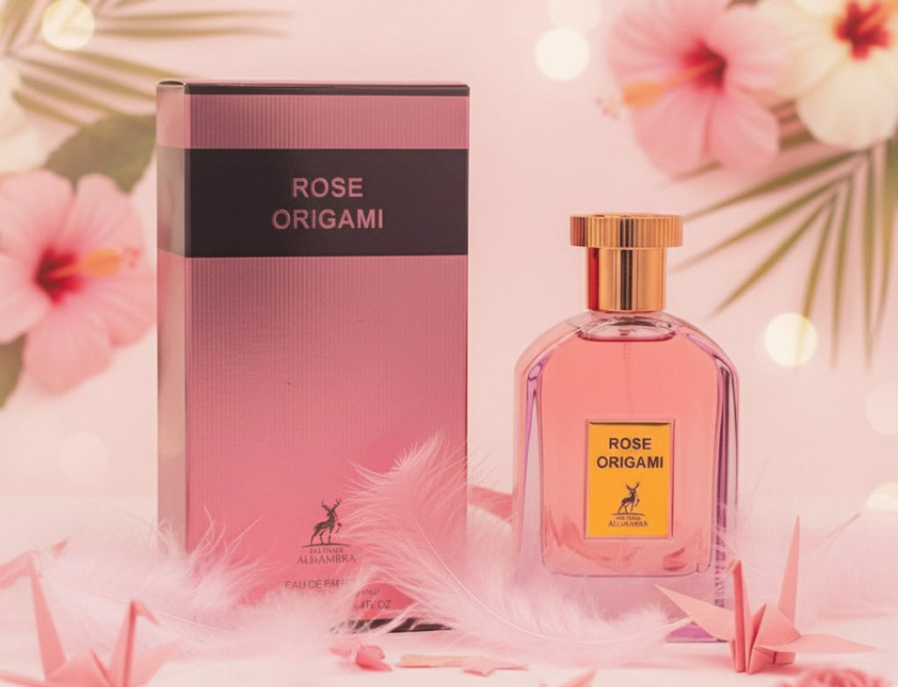 Maison Alhambra Rose Origami — Pink perfume bottle and box with gold accents, surrounded by pink origami cranes and feathers, with soft pink hibiscus flowers and bokeh lights in the background.