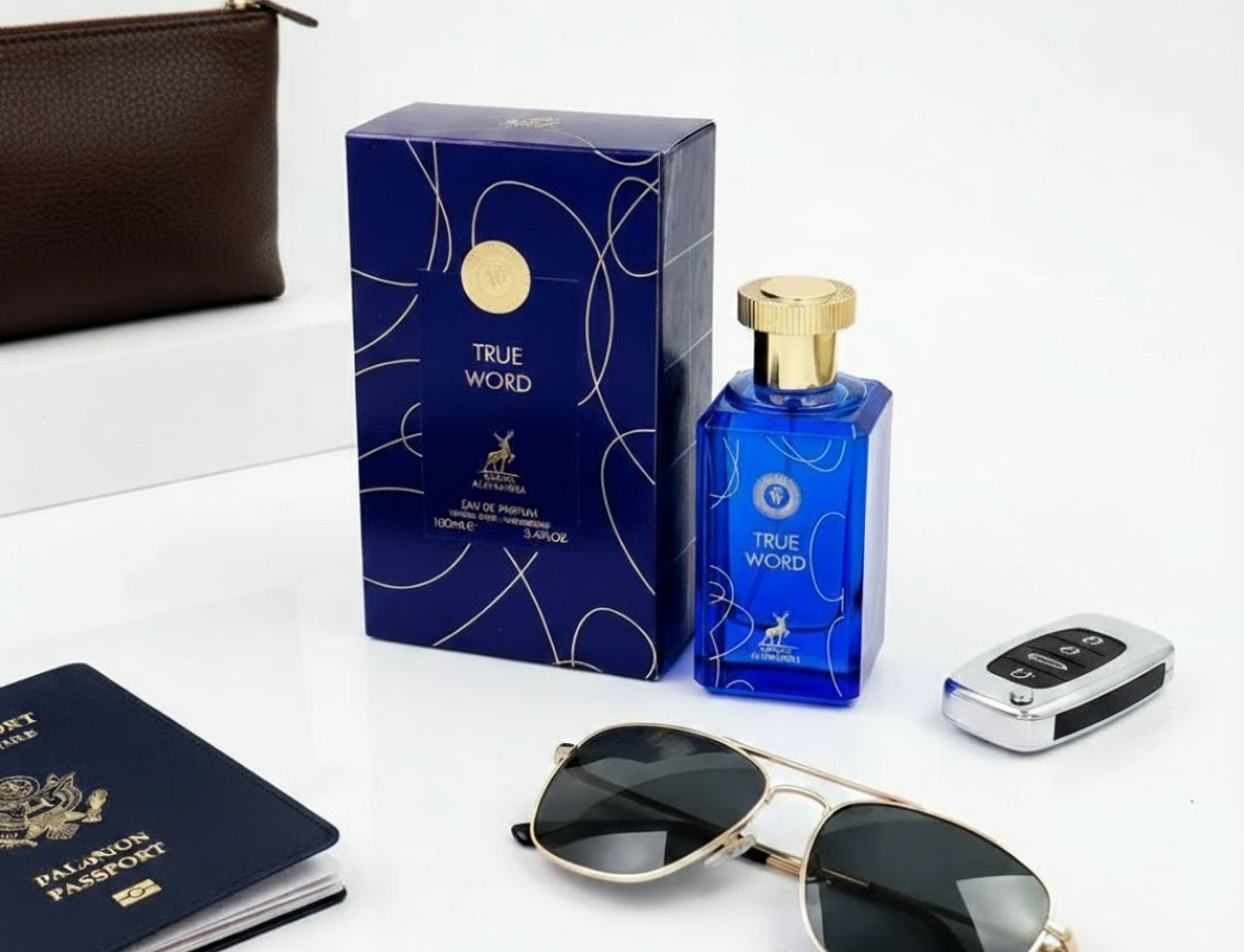 Maison Alhambra True Word by Maison Alhambra 100ml EDP — Blue perfume bottle with gold cap, matching blue box with gold swirls, sunglasses, passport, and car key fob on a white surface.