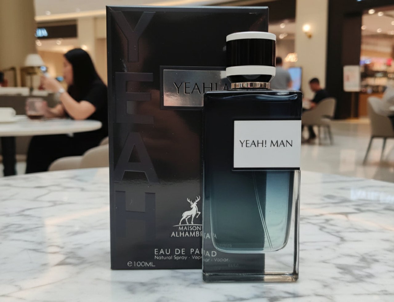 Maison Alhambra Yeah by Maison Alhambra 100ml EDP Men — Black box and glass bottle of fragrance with white label 'YEAH! MAN' on a marble surface, blurred background of a shopping mall.