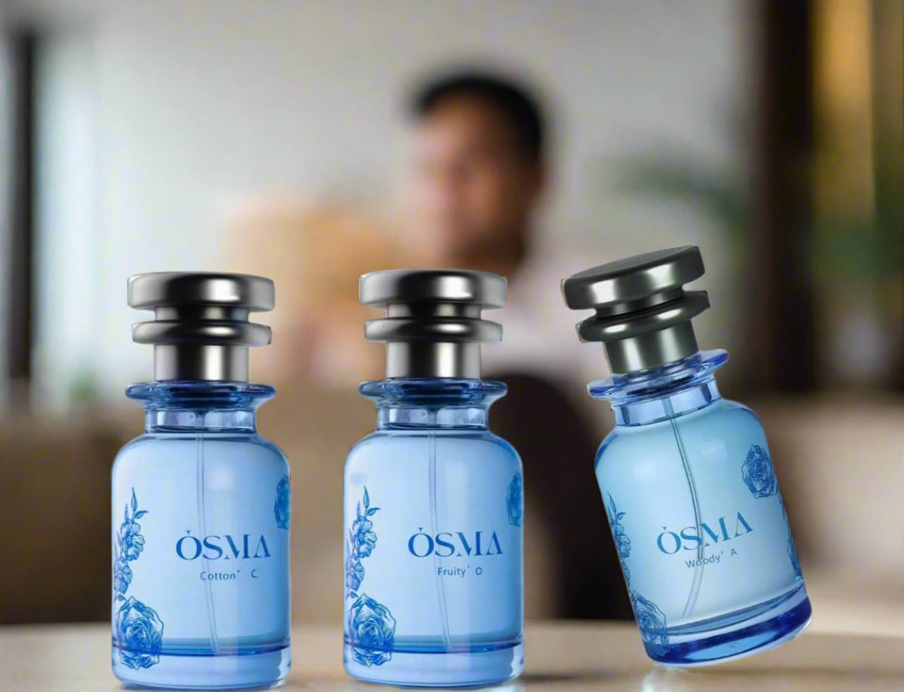 Osma Amber A Amber & Leather 75ml EDP — Tiga botol minyak wangi kaca biru bertutup perak, dihiasi reka bentuk bunga biru, disusun di atas permukaan berwarna cerah dengan orang kabur di latar belakang.