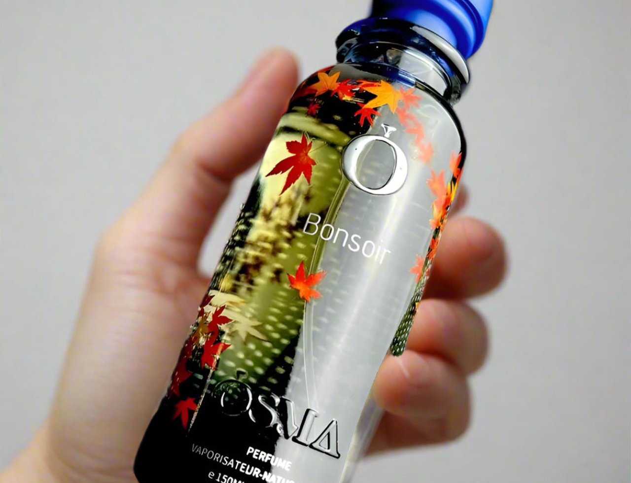 Osma Osma Bonsoir Cedarwood Warmth 150ml EDP — A hand holding a clear perfume bottle with a blue cap, decorated with red and orange maple leaves and a silver 'O' logo.