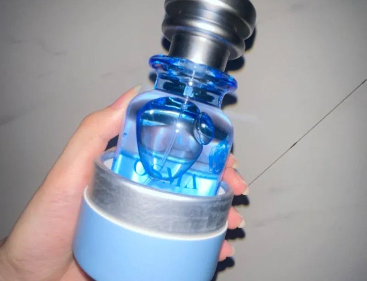 Osma Flowers W Osma Sandalwood 75ml — Hand holding a blue glass perfume bottle with a silver cap and a light blue base, against a grey tiled background.