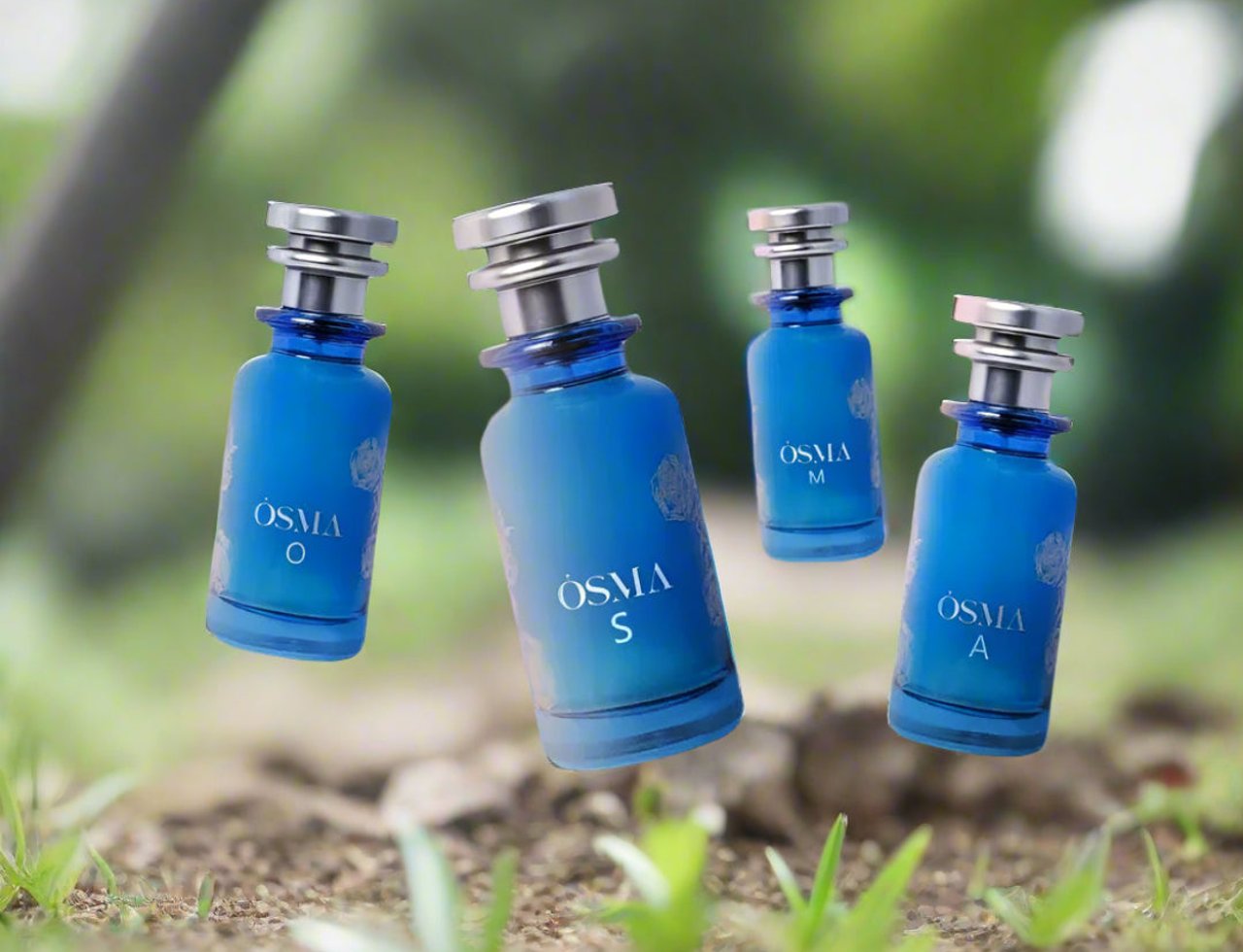 Osma Osma M Pineapple & Vanilla 150ml EDP — Four blue glass perfume bottles with silver caps floating in a natural outdoor setting with blurred green foliage and soil in the foreground.