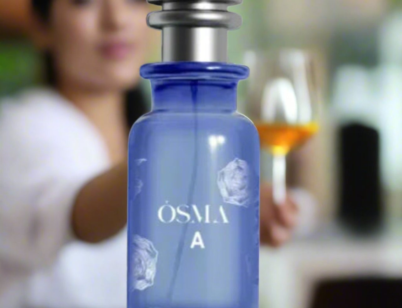 Osma Osma A Tropical & Soft 150ml EDP — Botol minyak wangi biru dengan penutup perak, ada corak bunga dan tulisan 'OSMA A' di hadapan. Wanita kabur pegang gelas wain di belakang.