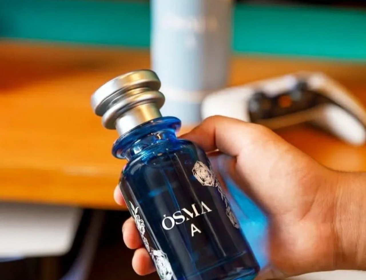 Osma Osma A Tropical & Soft 150ml EDP — Blue glass perfume bottle with silver cap held in hand, with a blurred background of a gaming controller and a light blue cylinder.