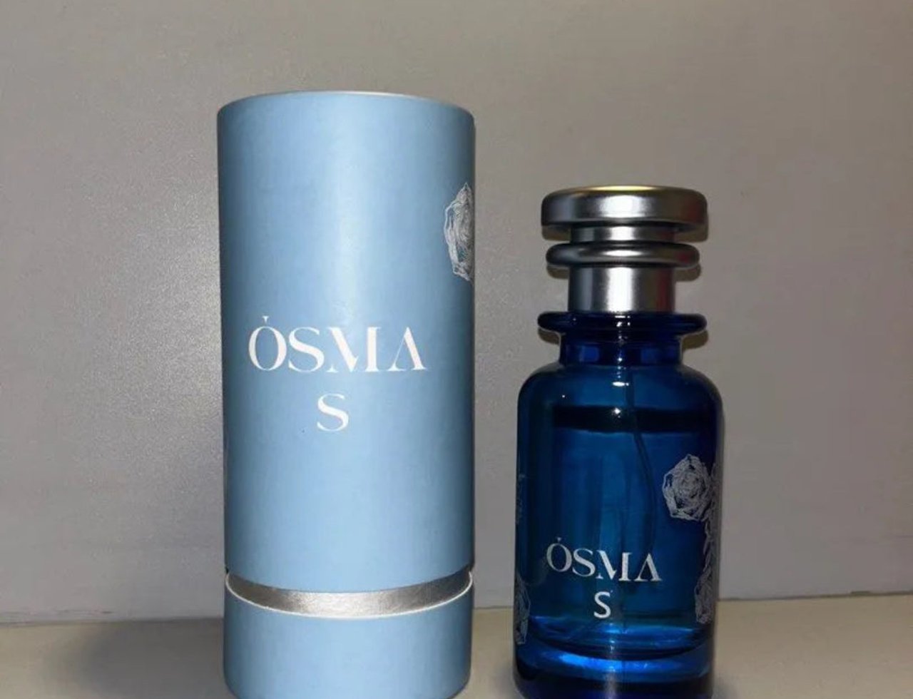 Osma Osma S – Pepper & Sandalwood EDP 150 ml — Botol minyak wangi kaca biru bertutup perak di sebelah kotak silindernya yang biru muda, kedua-duanya mempunyai tulisan putih dan grafik bunga.