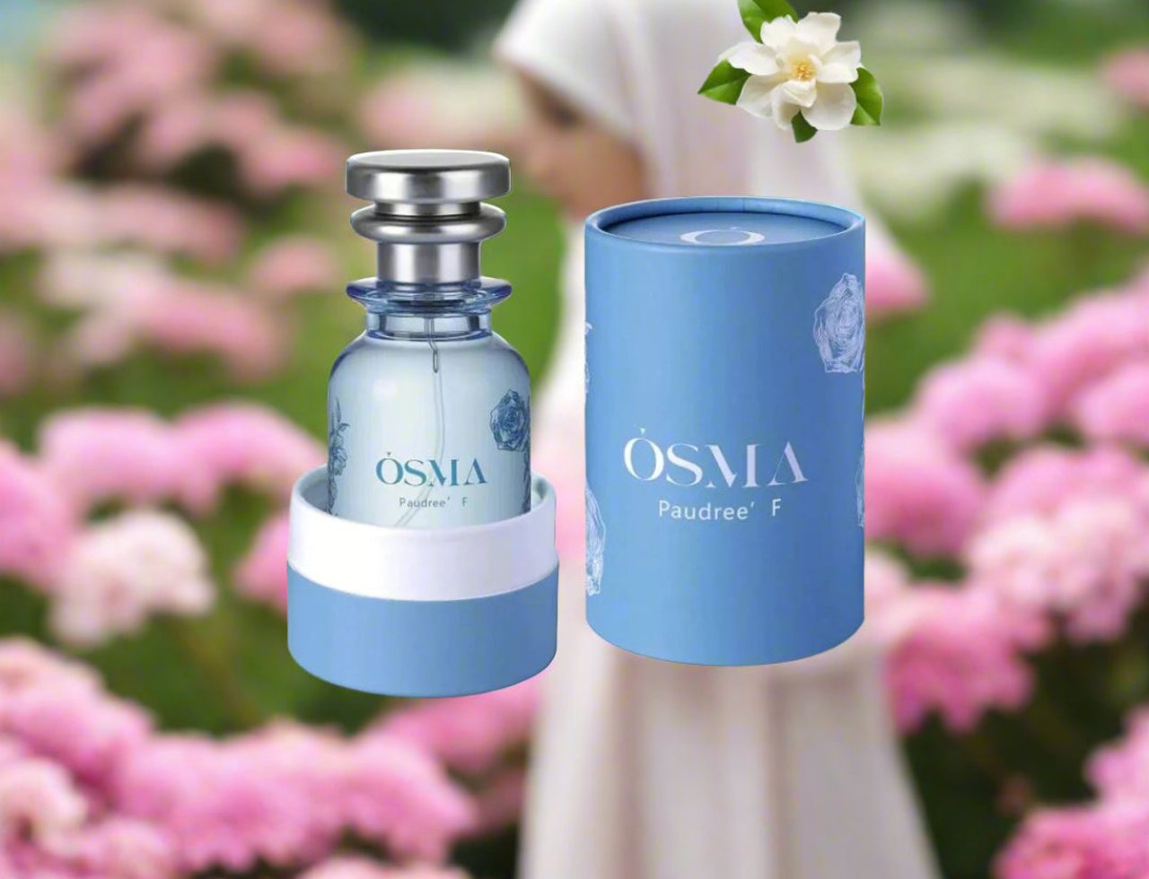 Osma Paudree F Roses & Musk 75ml EDP — A clear glass perfume bottle with a silver cap and blue accents, next to its blue cylindrical box, with a white flower and blurred pink flowers in the background.