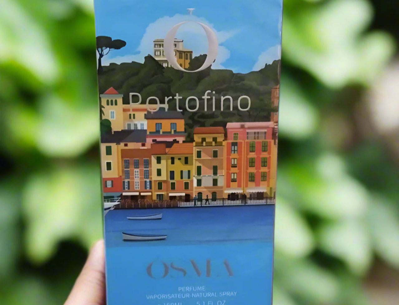 Osma Osma Portofino Summer Purity 150ml EDP — A hand holds a perfume box featuring a colorful illustration of Portofino, Italy, with buildings by the sea and boats.