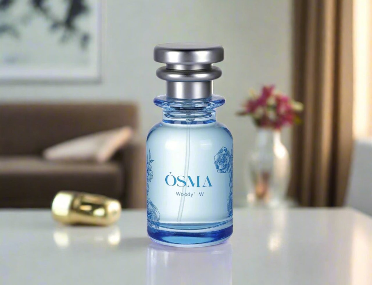 Osma Woody W Bergamot Leather 75ml EDP — Botol minyak wangi biru muda dengan corak bunga ros biru dan penutup perak, diletakkan di atas meja putih dengan penutup emas di sebelahnya. Latar belakang menunjukkan suasana ruang tamu yang kabur.