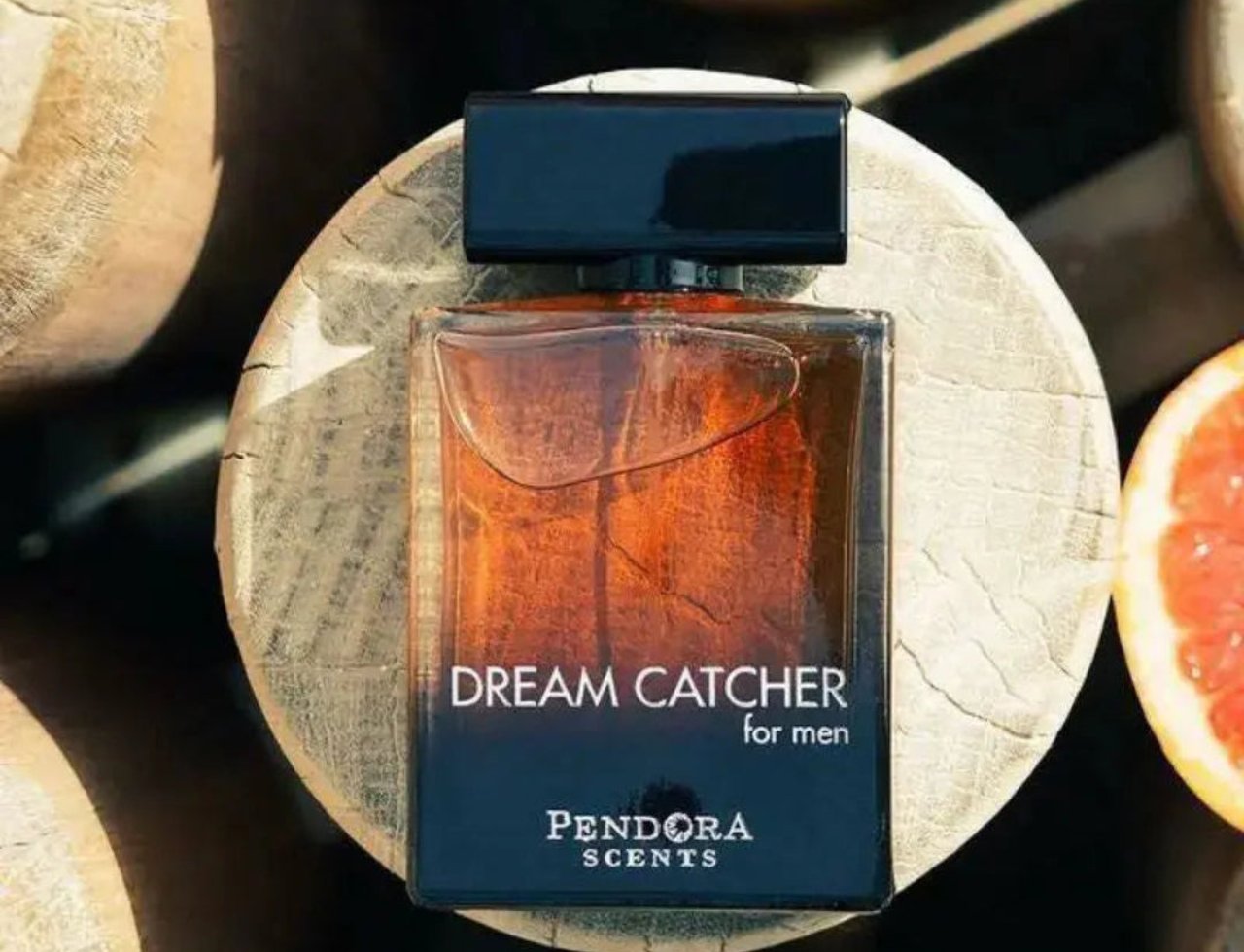 Paris Corner Dream Catcher by Paris Corner Pendora 100ml EDP — Botol minyak wangi kaca perang berbentuk segi empat dengan penutup hitam, diletakkan di atas tapak bulat kayu, dengan hirisan limau gedang kelihatan di sebelah kanan.