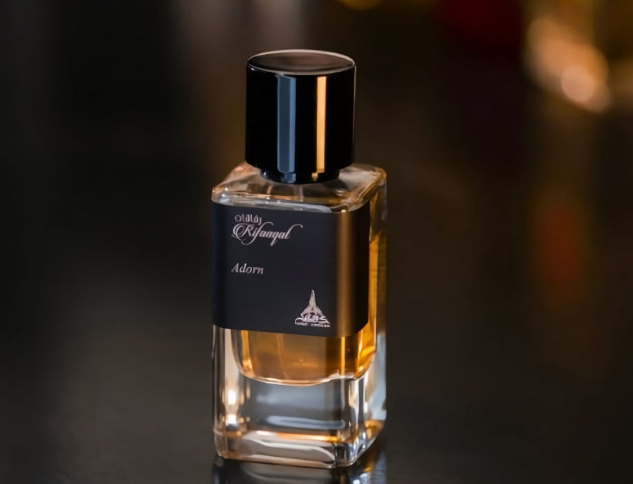 Paris Corner Rifaaqat Adorn by Paris Corner 85ml EDP — A clear glass perfume bottle with amber liquid, black cap, and a black label featuring Arabic and English text and a small building illustration, set against a dark, blurred background.
