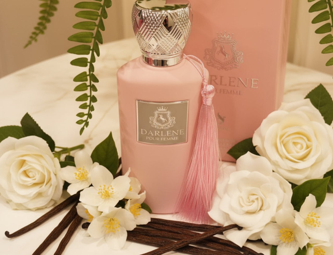 Volare Darlene Pour Femme by Volare – 100 ml untuk Wanita, botol merah jambu dengan penutup perak, dikelilingi bunga mawar putih, bunga melur, dan kacang vanila, dengan kotak merah jambu di latar belakang.
