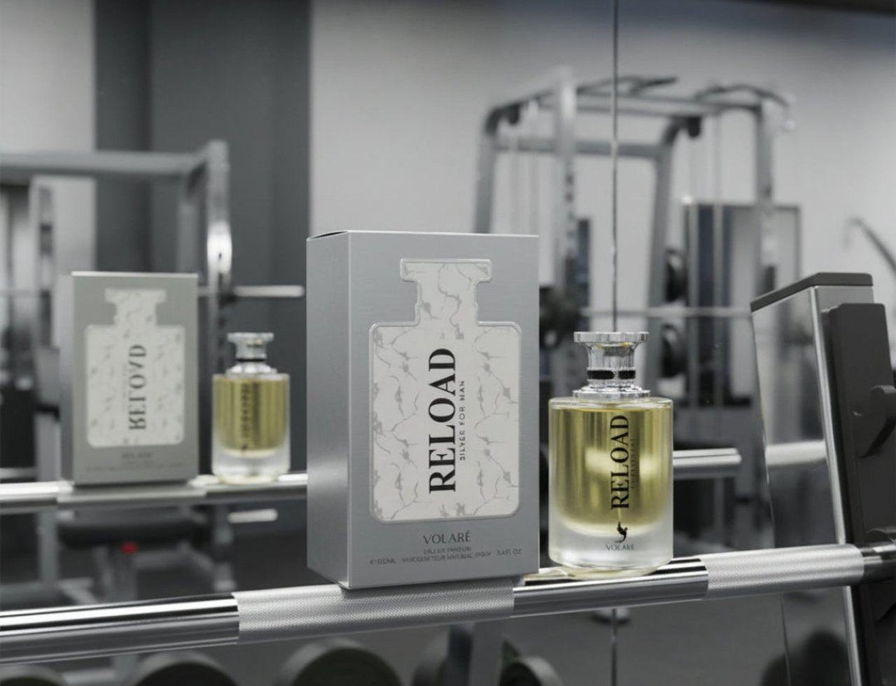 Botol dan kotak minyak wangi Volare Reload Silver by Volare – Man 100 ml for Him dipamerkan di atas barbel gimnasium, dengan kelengkapan gimnasium di latar belakang.