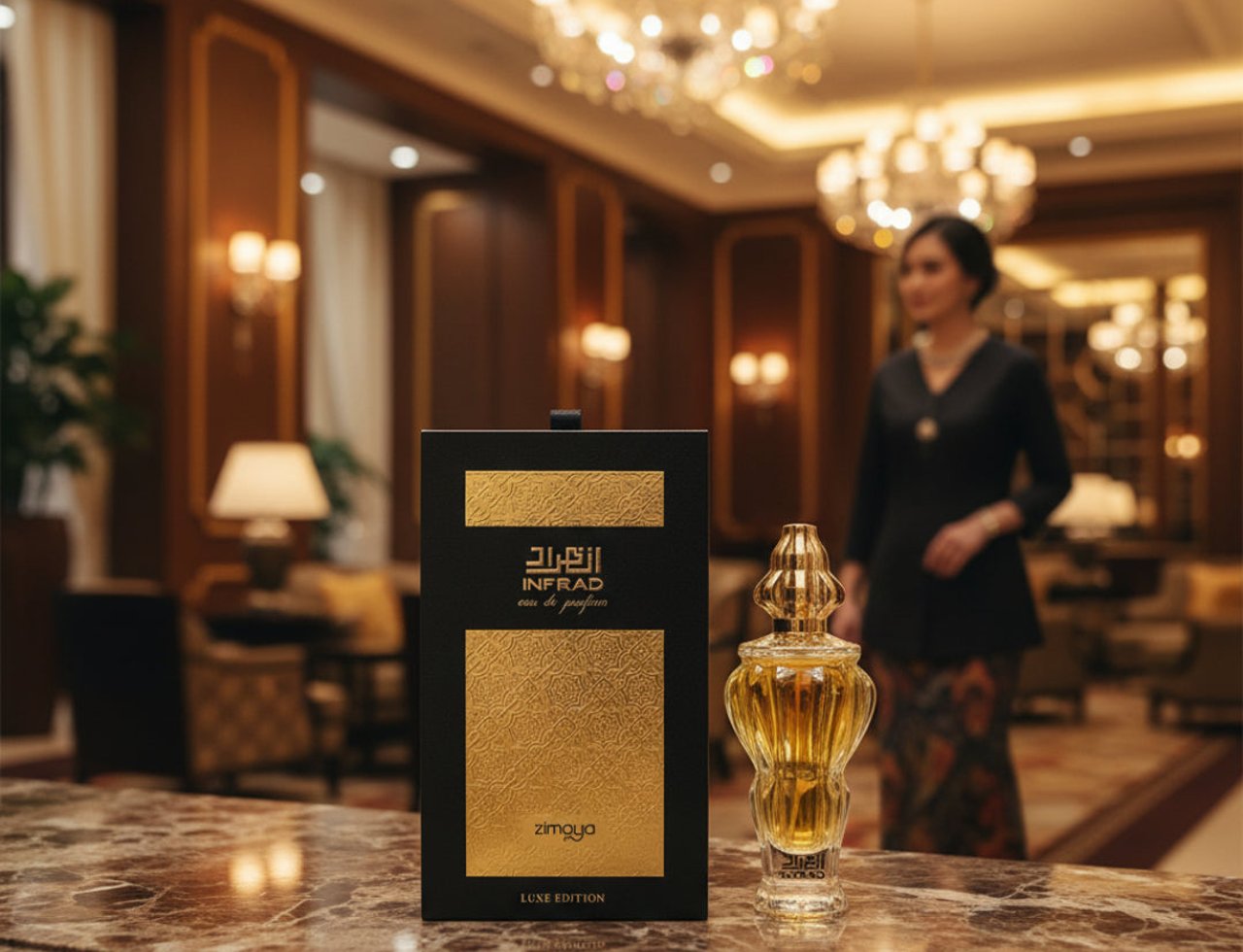 Zimaya Infrad Luxe by Zimaya 100ml EDP untuk Wanita — Kotak wangian hitam dan emas dengan tulisan Arab dan panel bertekstur emas, bersebelahan botol wangian kaca jernih berpenutup emas berukir, di atas meja marmar, dengan wanita di latar belakang.