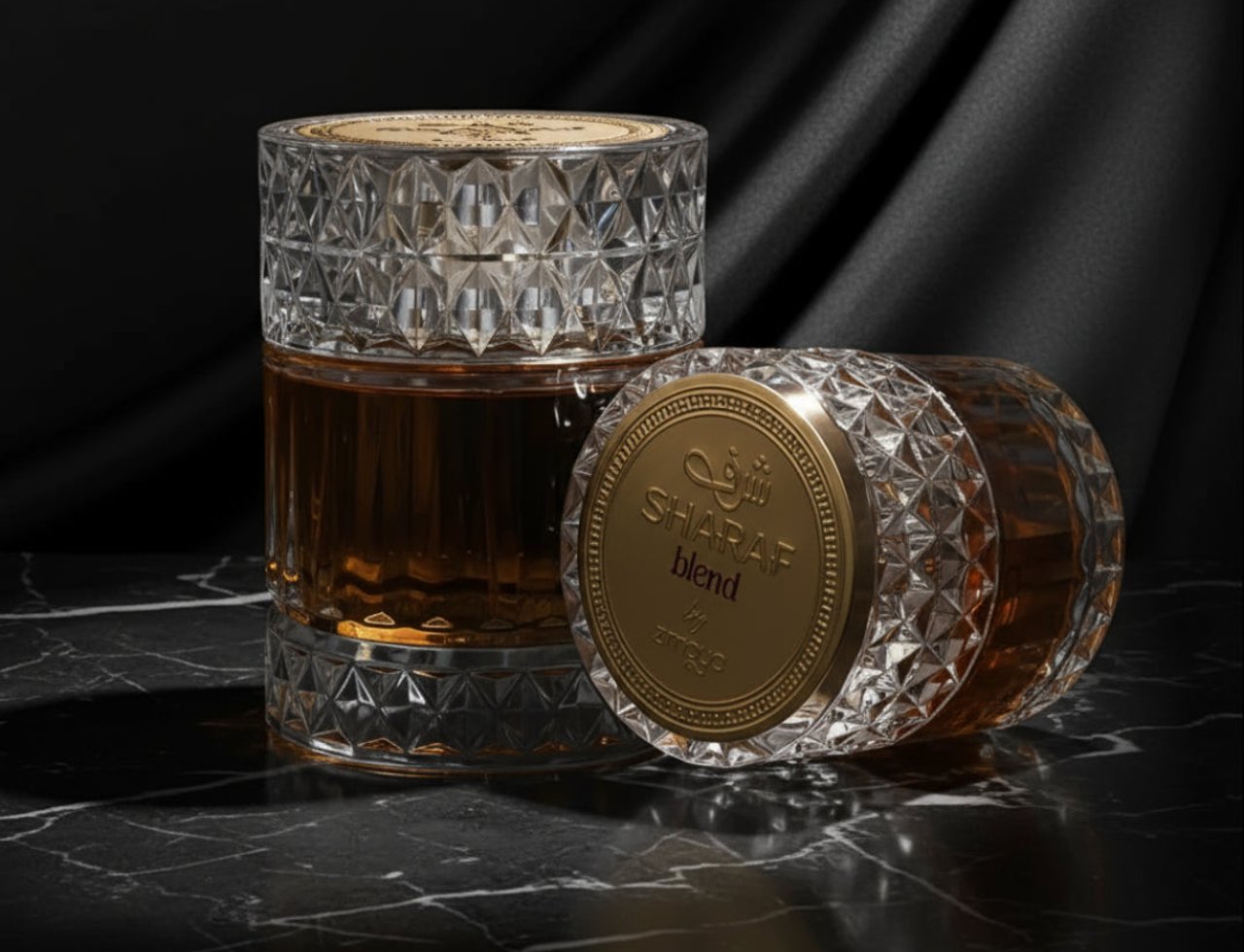 Zimaya Sharaf Blend by Zimaya 100ml Extrait de Parfum — Two crystal perfume bottles with gold caps, one upright and one lying down, on a black marble surface with a draped black fabric background.
