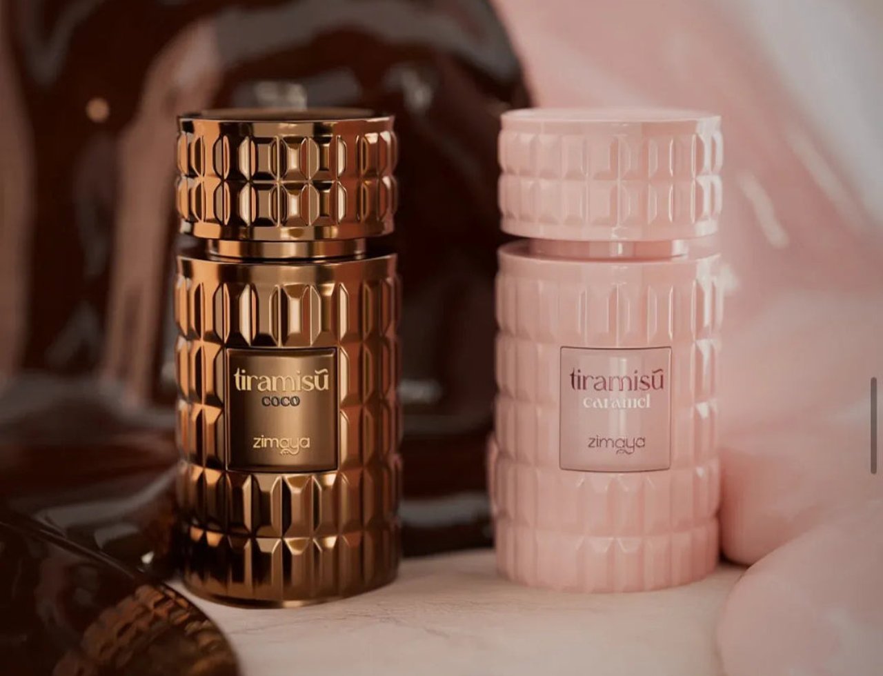 Zimaya Tiramisu Caramel By Zimaya 100 ml EDP — Two perfume bottles, one gold with 'tiramisu coco' label and one pink with 'tiramisu caramel' label, on a light surface with a blurred chocolate and pink background.