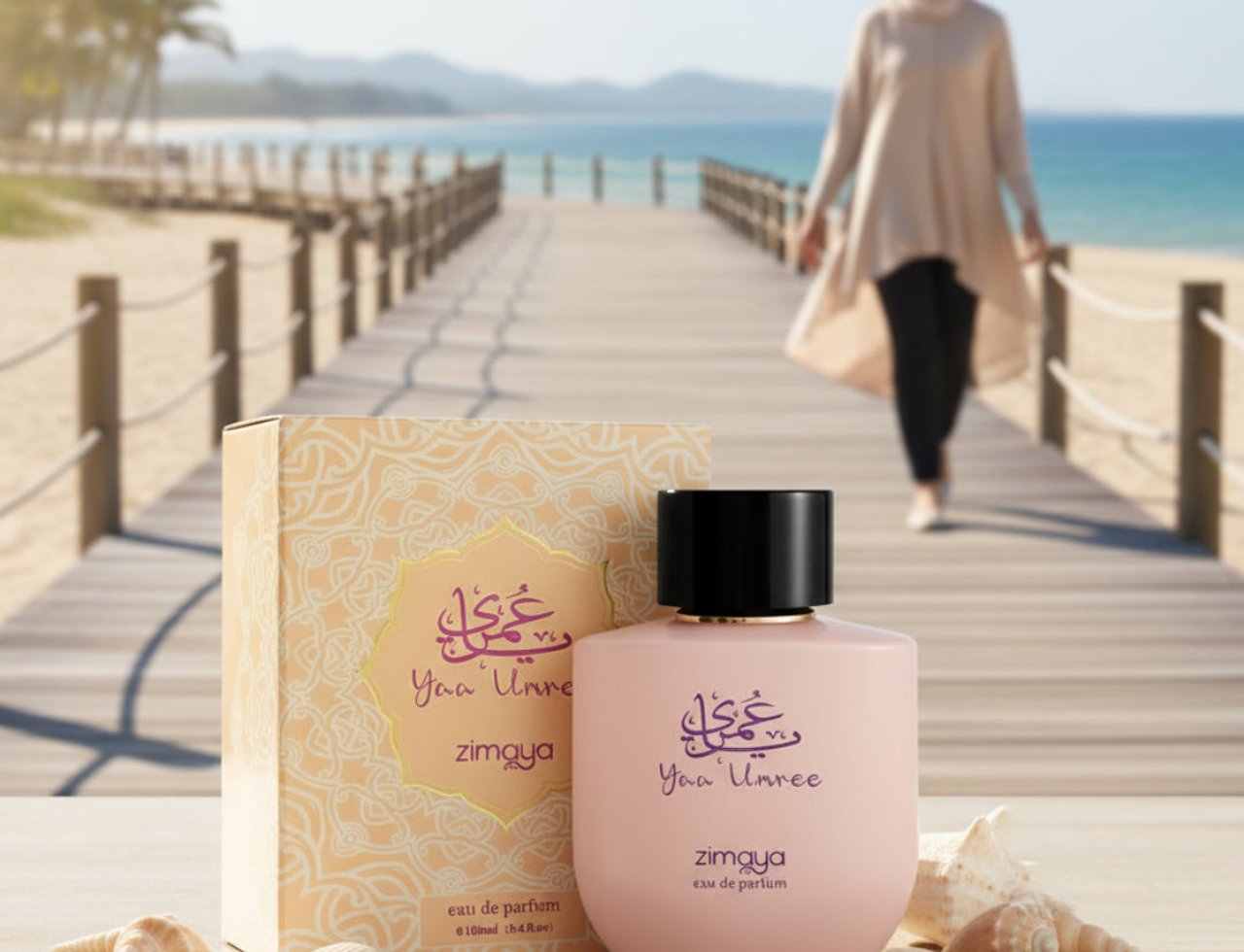 Zimaya Yaa Umree by Zimaya EDP 100 ml Women — Botol dan kotak minyak wangi merah jambu di atas permukaan kayu dengan cengkerang laut, laluan pejalan kaki dan pantai di latar belakang.