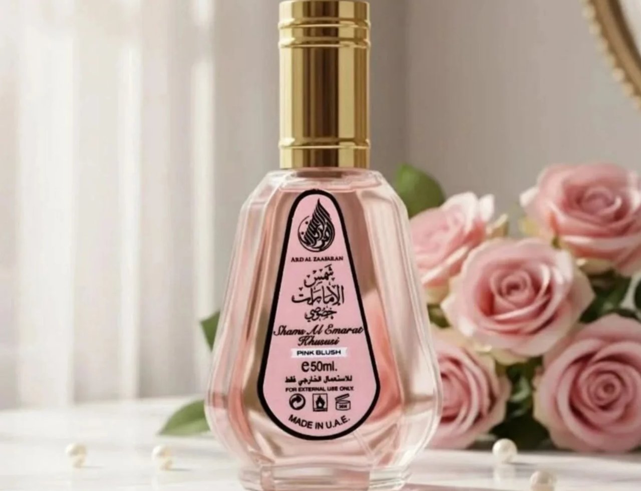 Ard Al Zaafaran Pink Blush Shams Al Emarat Khususi 50ml — A clear glass perfume bottle with pink liquid and a gold cap, set against a backdrop of pink roses and soft lighting.
