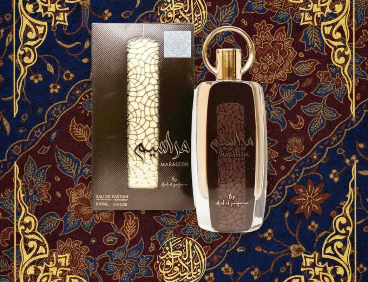 Ard Al Zaafaran Maraseem by Ard Al Zaafaran – 100 ml Unisex, menampilkan botol kaca coklat dengan corak berjalin emas dan penutup bulat emas, diletakkan di sebelah kotak kadbod coklat dengan corak berjalin putih. Latar belakang adalah kain bercorak bunga biru dan merah dengan hiasan tepi emas.
