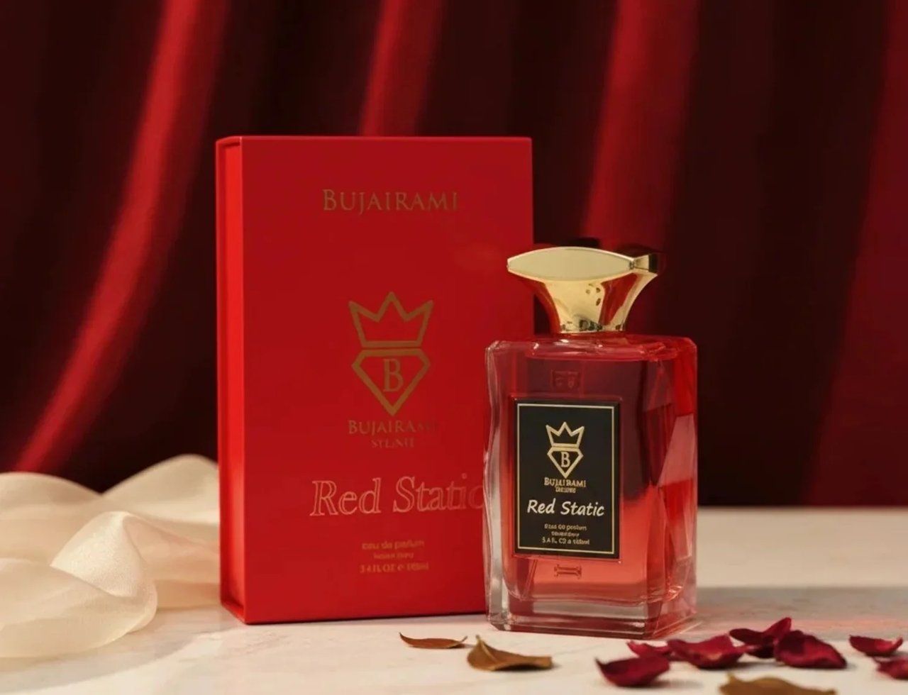 Bujairami Red Static by Bujairami Eau de Parfum 100ml — Red perfume bottle with gold cap next to its red box, set against a red draped background with scattered rose petals and sheer fabric.