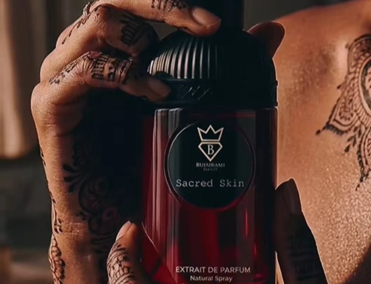 Bujairami Sacred Skin 100ml Extrait Unisex — Tangan berhiasan inai rumit memegang botol minyak wangi merah gelap bertutup hitam berjalur.