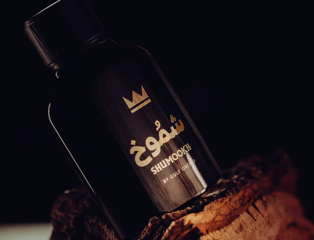 Gulf Orchid Shumookh Parfum 100ml — Botol wangian hitam legam dengan tulisan emas dan simbol mahkota, bersandar pada kayu bertekstur gelap dengan latar belakang gelap.