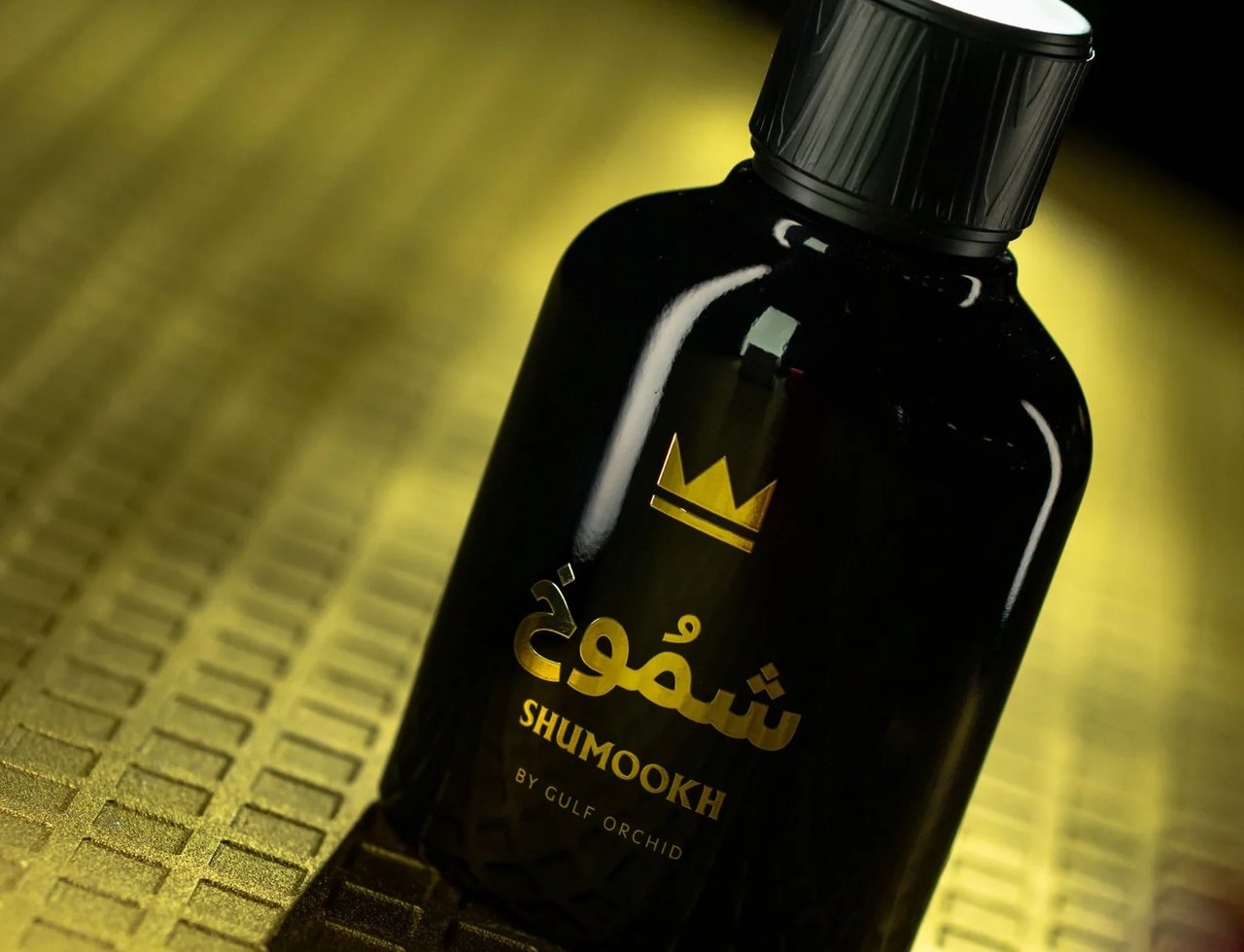 Gulf Orchid Shumookh Parfum 100ml by Gulf Orchid — A black perfume bottle with a gold crown logo and Arabic script, tilted on a textured gold surface. | Gulf Orchid Shumookh Parfum 100ml by Gulf Orchid — Botol minyak wangi hitam dengan logo mahkota emas dan tulisan Arab, condong di atas permukaan emas bertekstur.