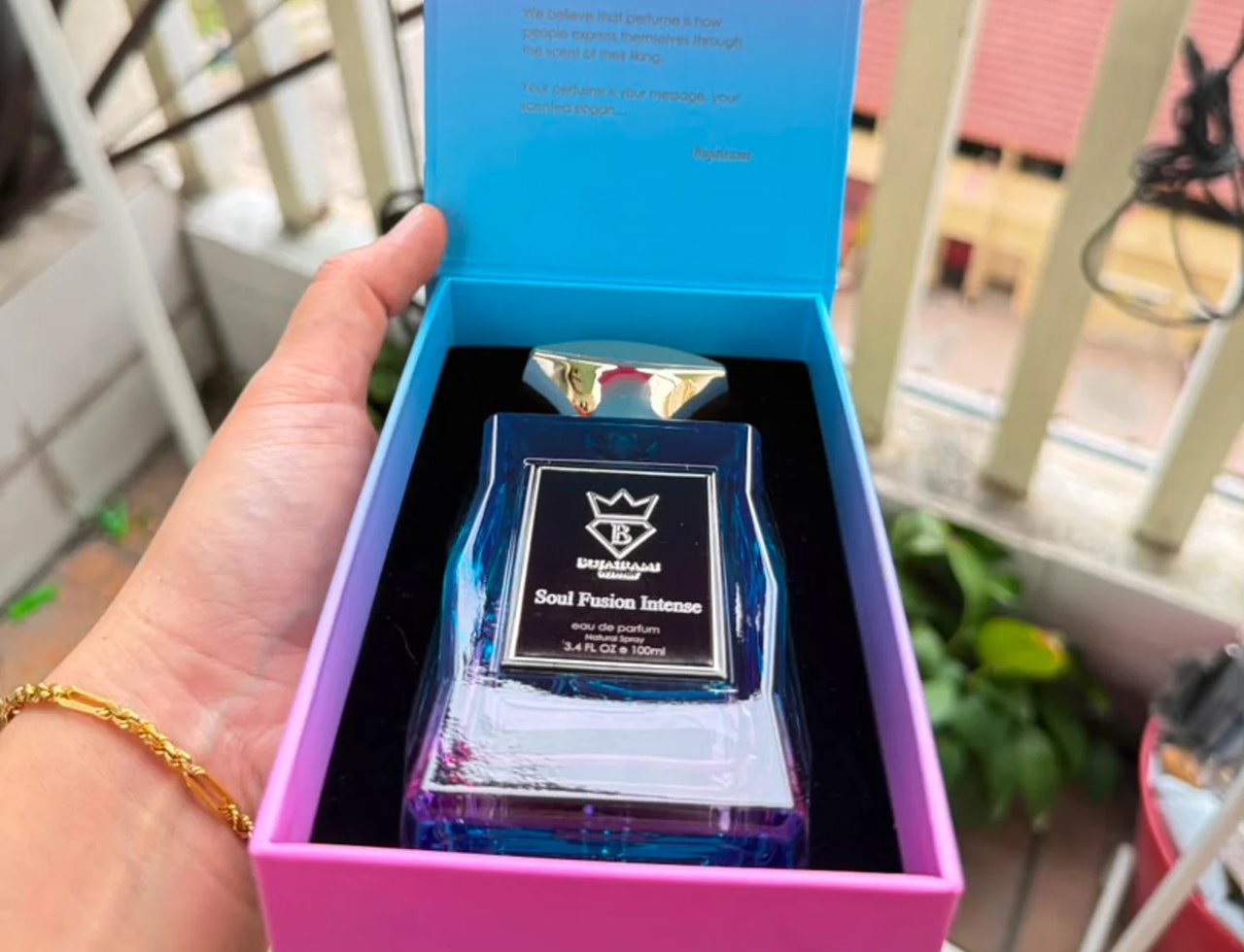 Bujairami Soul Fusion Intense — Tangan memegang botol minyak wangi kaca biru bertutup emas, dalam kotak gradien merah jambu dan biru, dengan tulisan di dalam penutup.