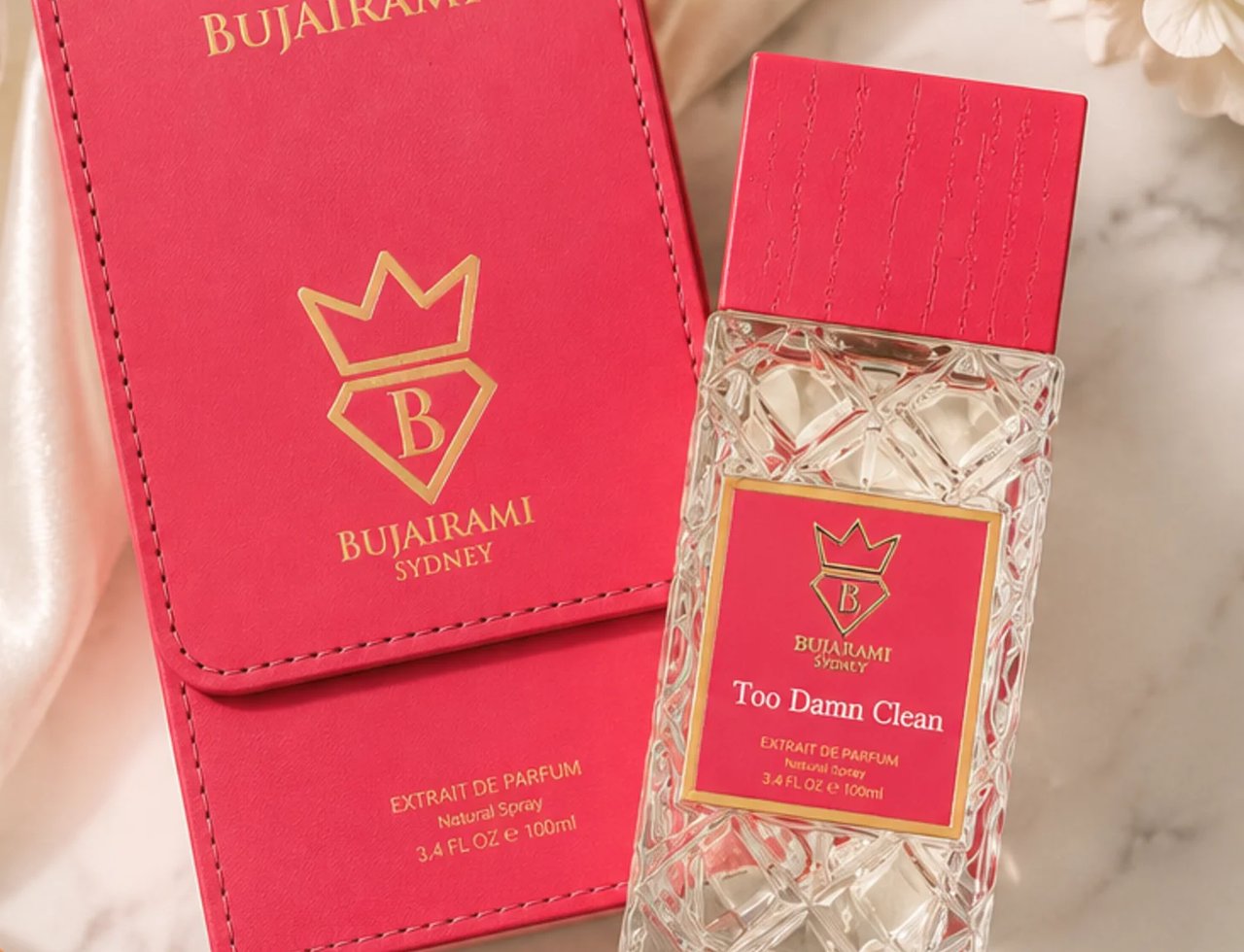 Bujairami Too Damn Clean — A clear glass perfume bottle with a textured red cap and a pink label featuring a gold crown and 'B' logo, placed next to its matching pink presentation box with gold lettering and logo.