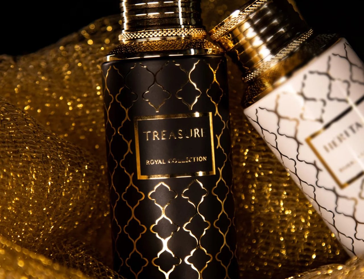 Maison Asrar Treasure by Maison Asrar 100ml EDP Unisex — Two ornate perfume bottles, one black with gold pattern and one white with gold pattern, nestled in shimmering gold fabric.