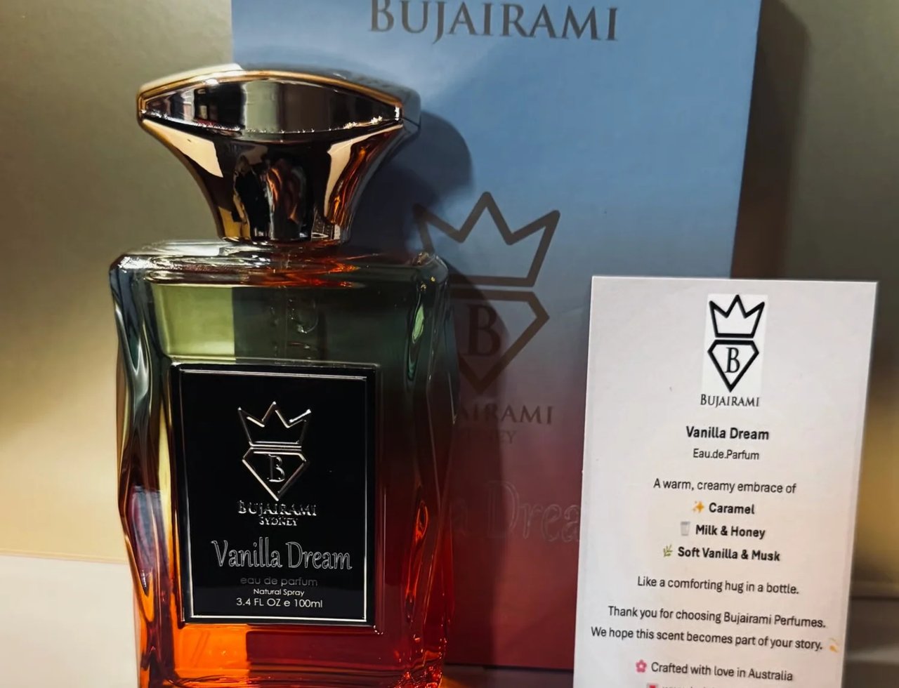 Bujairami Vanilla Dream Eau de Parfum — Botol minyak wangi kaca dengan cecair perangkap ke jernih, penutup emas, dan label hitam. Kotak biru muda dan kad maklumat produk di belakangnya.