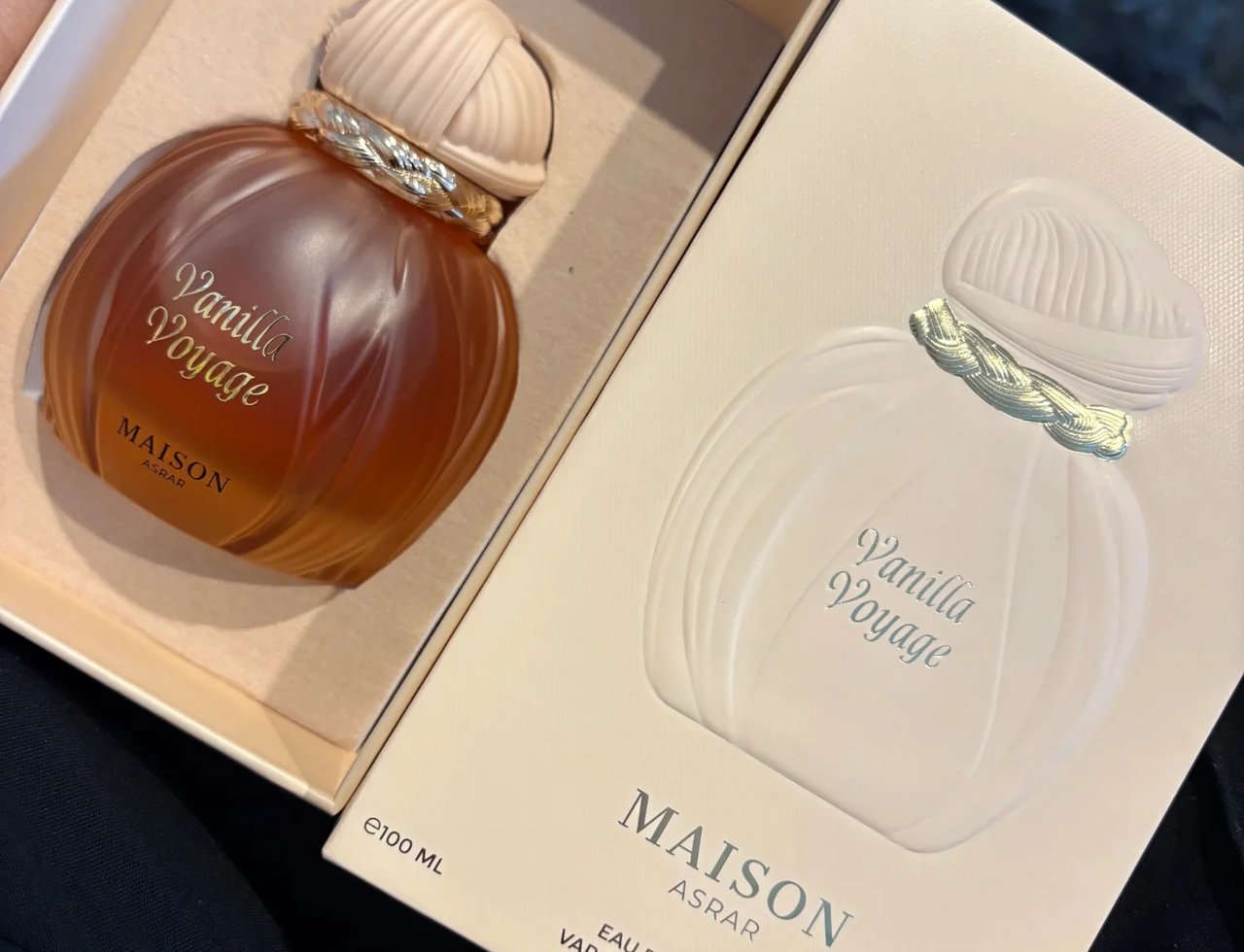 Maison Asrar Vanilla Voyage — Botol minyak wangi berwarna ambar dengan penutup bertekstur dan trim emas, diletakkan dalam kotak beige terbuka bersebelahan kotak produk sepadan dengan butiran timbul.