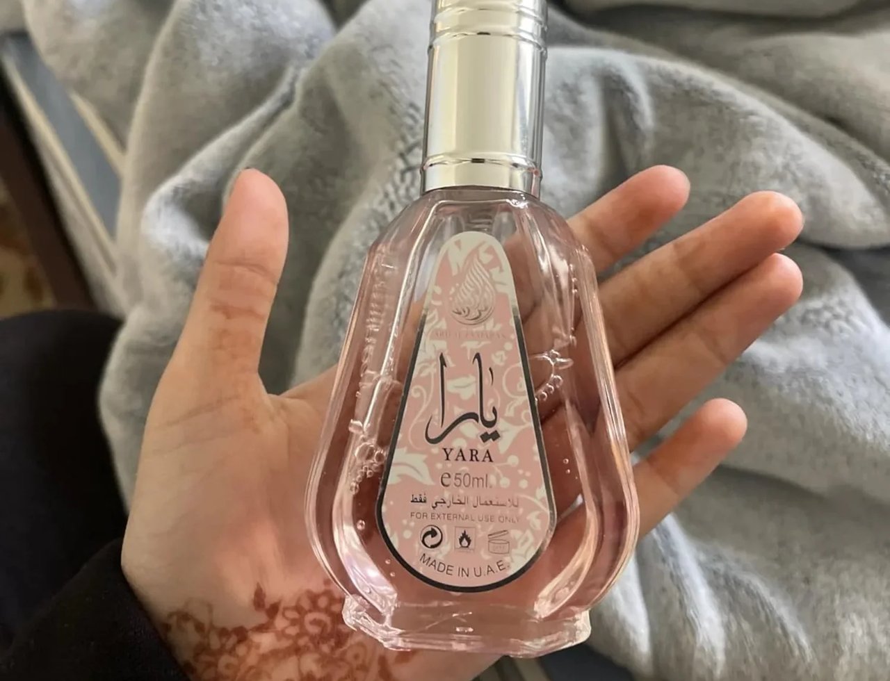 Ard Al Zaafaran Yara Eau De Parfum 50ml by Ard Al Zaafaran — Botol kaca jernih berisi cecair merah jambu dipegang tangan dengan corak inai di tapak tangan, latar belakang selimut kelabu
