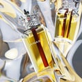 Lattafa Dynasty by Lattafa 100ml EDP Unisex — A clear glass perfume bottle with yellow liquid and a red rod inside, topped with a silver crown cap with red gems. The bottle is angled, with a second bottle blurred in the background, set against a shimmering, abstract silver and gold fabric.