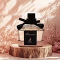 Maison Alhambra Florenza (Florence) 100ml — A clear glass perfume bottle with a black label featuring a deer silhouette and "Florenza" text, adorned with a black ribbon, sits on a wooden pedestal against a soft pink, textured background with dried flowers. | Maison Alhambra Florenza (Florence) 100ml — Botol minyak