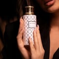 Maison Asrar Heritage by Maison Asrar 100ml EDP Unisex — Tangan wanita memegang botol minyak wangi corak putih dan emas dengan penutup emas, berlatar belakang gelap.