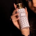 Maison Asrar Heritage by Maison Asrar 100ml EDP Unisex — Tangan memegang botol minyak wangi putih bercorak emas dan penutup emas, berlatar belakang gelap.