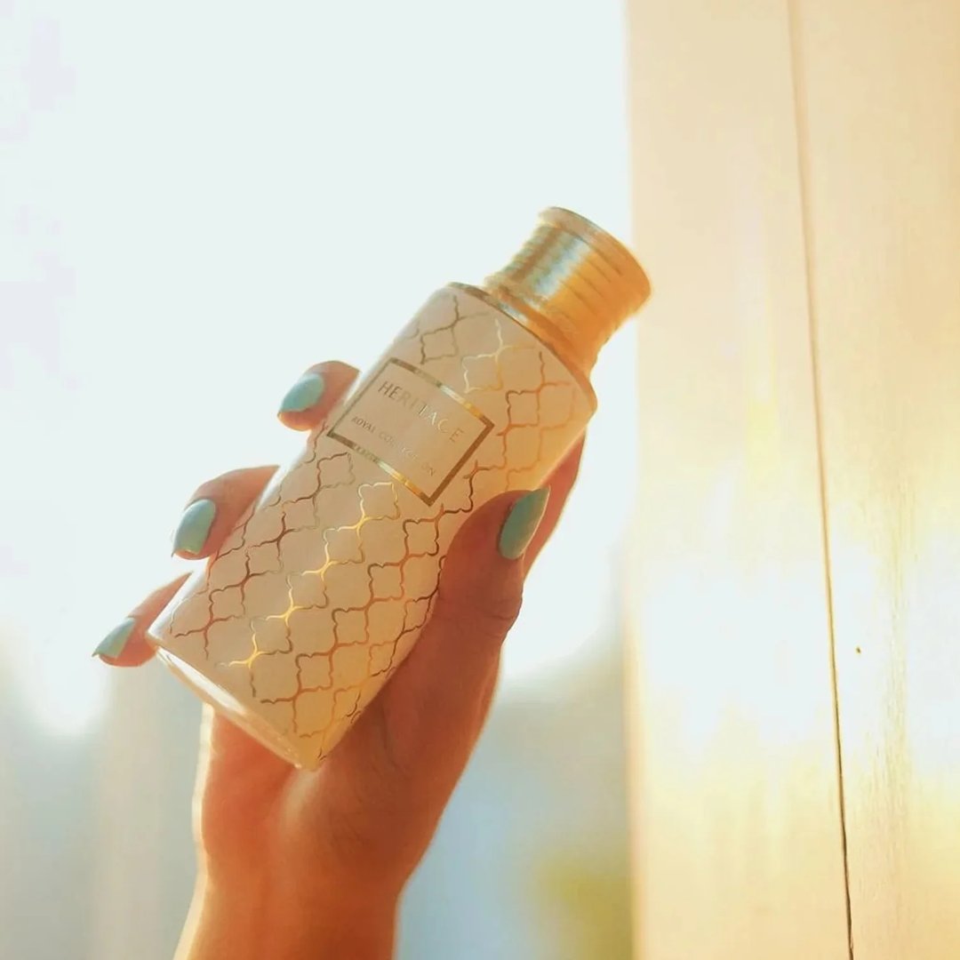 Maison Asrar Heritage by Maison Asrar 100ml EDP Unisex — Tangan memegang botol putih bercorak geometri emas dan penutup emas, berlatar belakang cerah disinari matahari.