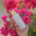 Maison Asrar Heritage by Maison Asrar 100ml EDP Unisex — Tangan dengan kuku biru muda memegang botol minyak wangi putih bercorak geometri emas dan penutup emas, dikelilingi bunga kertas merah jambu terang.