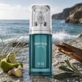French Avenue Imperial Ocean 100ml for Men — A tall teal glass perfume bottle with a silver ornate cap, set on a rocky beach with ocean waves in the background. Lime, ginger, and herbs are visible.