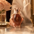 Zimaya Kibria Glory by Zimaya 100ml Wanita — Tangan wanita memegang botol minyak wangi emas ros bertutup mahkota, bunga ros merah jambu lembut dalam pasu kaca di latar belakang.