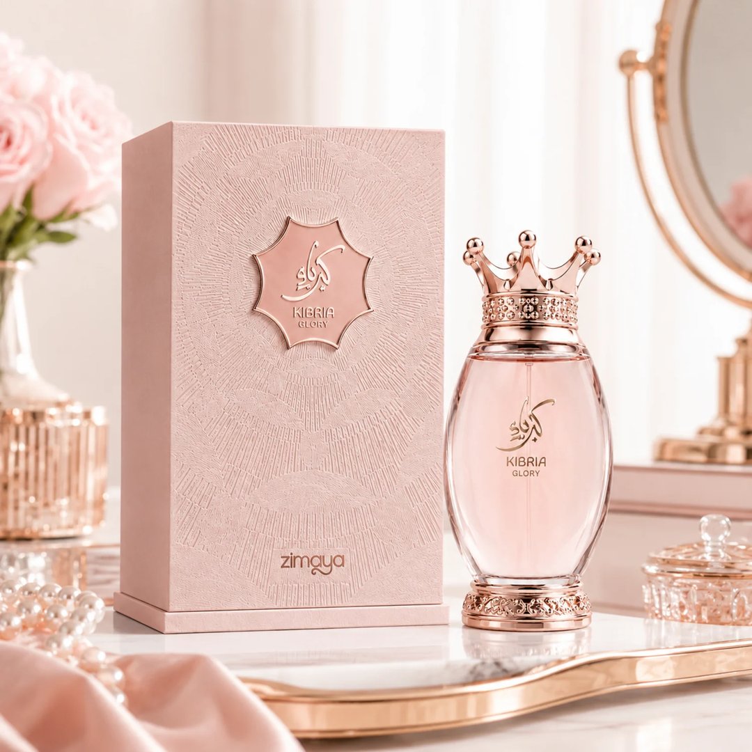 Zimaya Kibria Glory by Zimaya 100ml Wanita — Kotak merah jambu bertekstur dan botol minyak wangi kaca bujur dengan penutup berbentuk mahkota, diletakkan di atas permukaan marmar bersama mawar merah jambu dan mutiara.