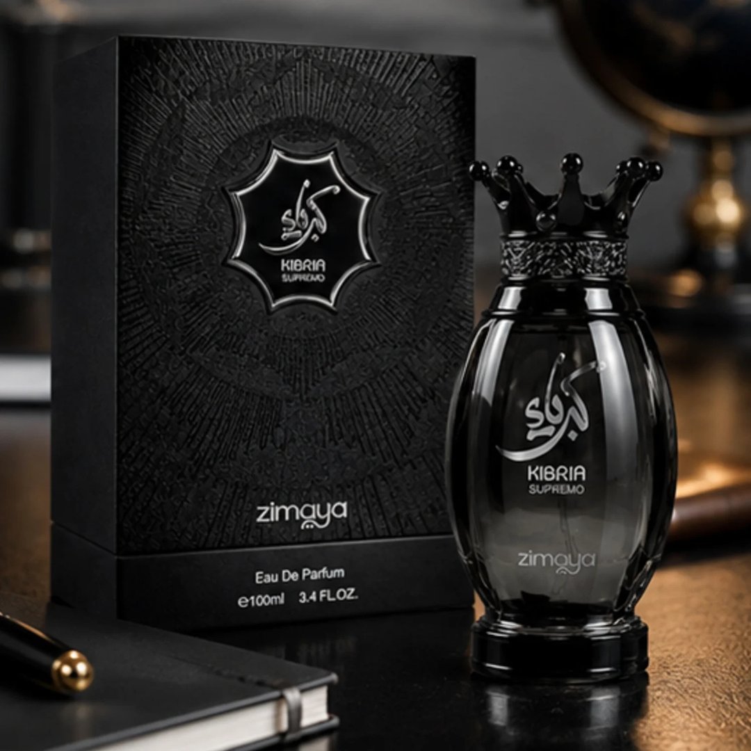 Zimaya Kibria Supremo oleh Zimaya 100ml Man — Kotak hitam bertekstur dan botol minyak wangi hitam bertutup mahkota dengan kaligrafi Arab perak dan teks di permukaan gelap yang memantul, dengan buku nota dan pen di latar depan.